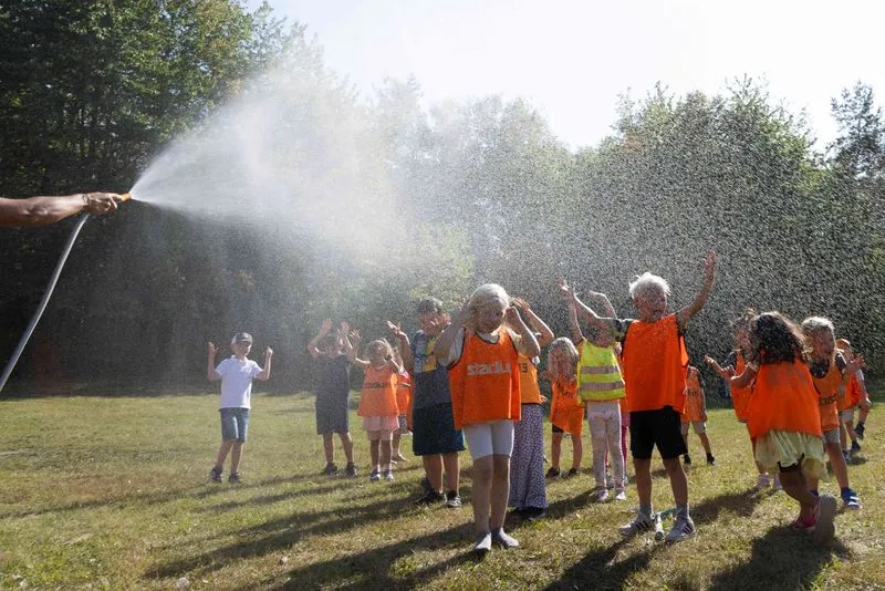 En grupp glada barn i orangea västar leker och blir nedstänkta av en vattenslang på en gräsmatta en solig dag, med skog i bakgrunden.