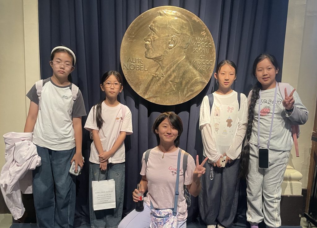 En grupp barn och en vuxen ledare poserar och ler framför en stor gyllene medalj med Alfred Nobels porträtt, troligen på Nobelmuseet.