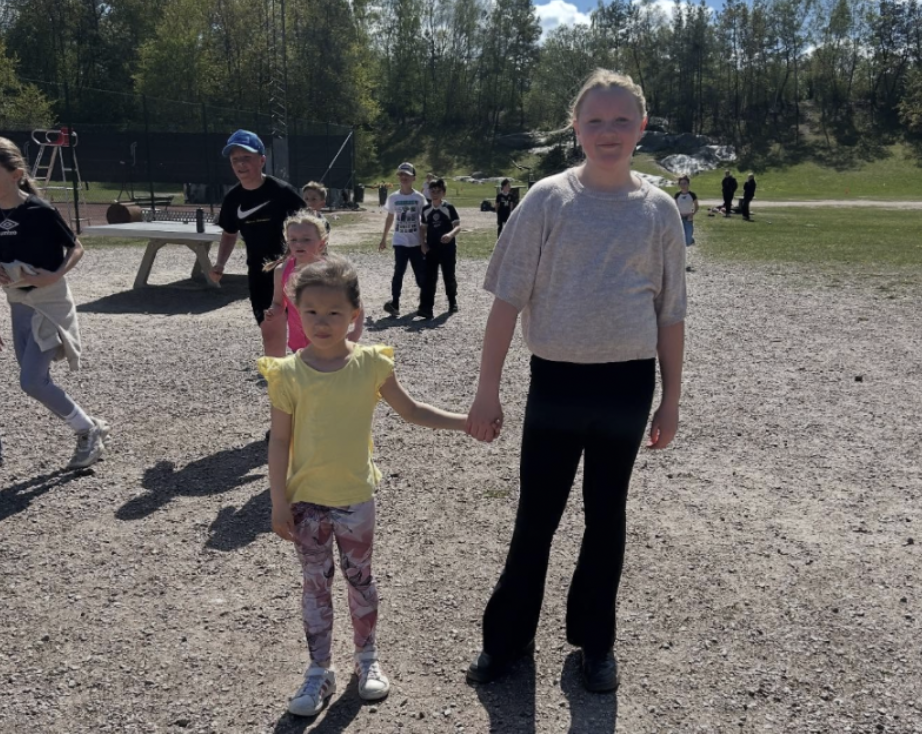 "En flicka och en tonåring håller varandra i handen utomhus på en grusplan en solig dag."