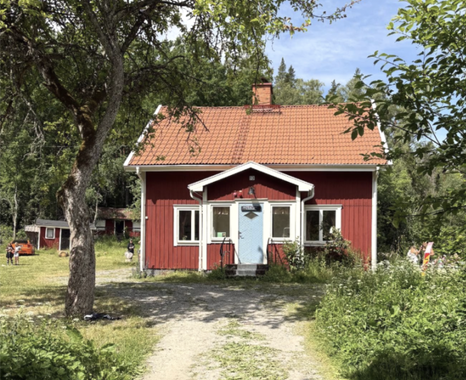 "Ett rött hus med vita knutar och ett rött tegeltak, omgivet av träd och grönska."